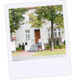 zdjęcie głównego wejścia do zabytkowej białej willi z widocznymi schodami i poręczami. Obok wysokie drzewa