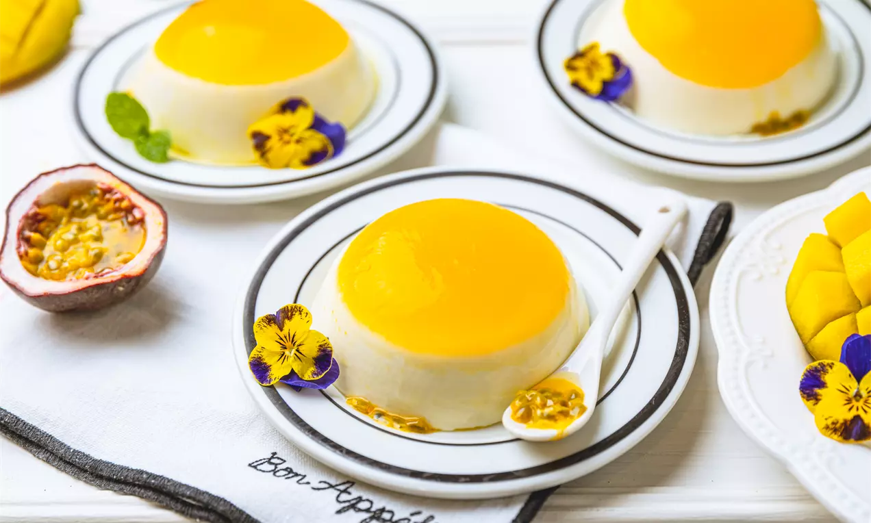 Na białym talerzyku panna cotta o gładkiej, białej bazie z intensywnie żółtym musem, ozdobiona jadalnymi kwiatami i owocem marakui