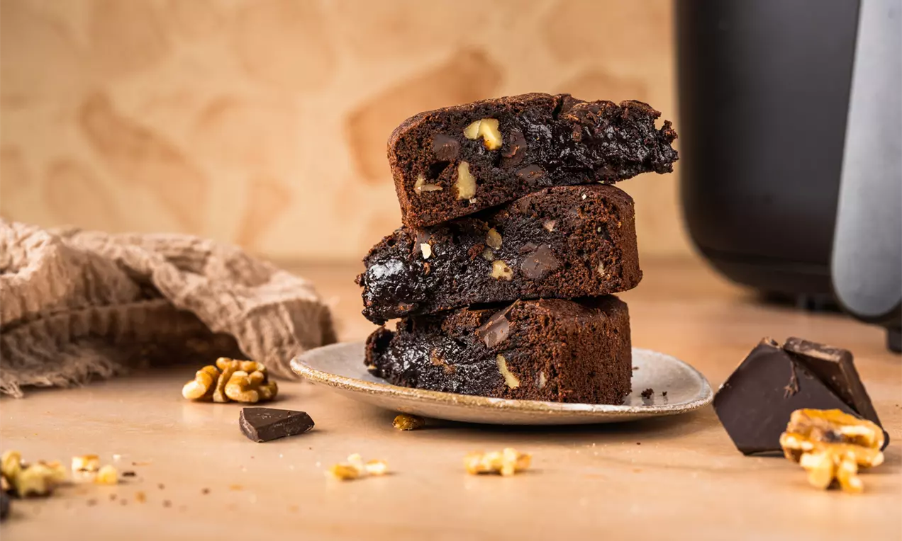 Trzy kawałki wilgotnego brownie z wyraźnymi kawałkami orzechów i czekolady, ułożone na jasnym talerzu, obok orzechów i czekolady