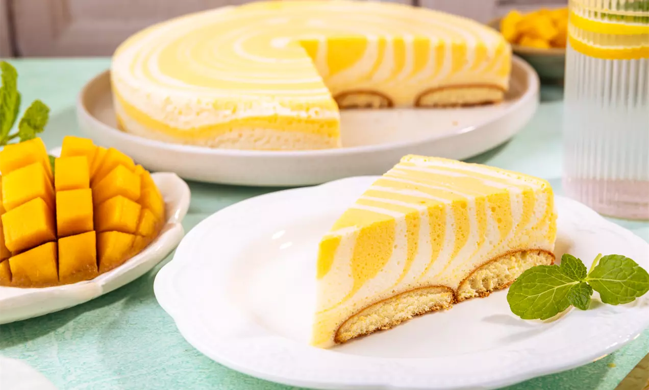 Delikatne ciasto na spodzie biszkoptowym z mango-jogurtową pianką o marmurkowym wzorze w odcieniach żółci i bieli, ozdobione listkiem mięty