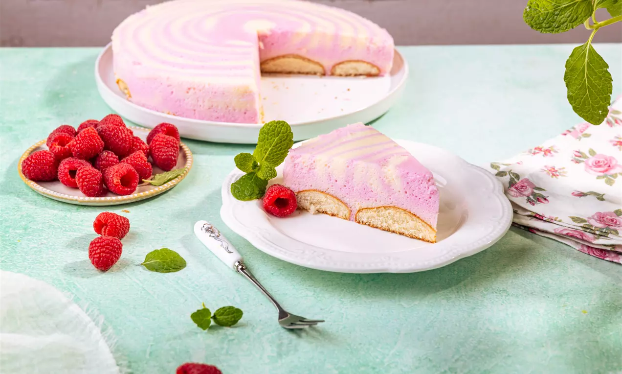 Ciasto w pastelowych odcieniach różu i bieli, z warstwą biszkoptów, na białym talerzu z malinami i listkiem mięty na dekoracyjnym tle