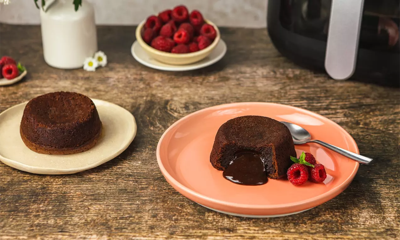 Czekoladowe lava cake z płynnym wnętrzem na brzoskwiniowym talerzu, ozdobione malinami i listkiem mięty, obok srebrna łyżeczka