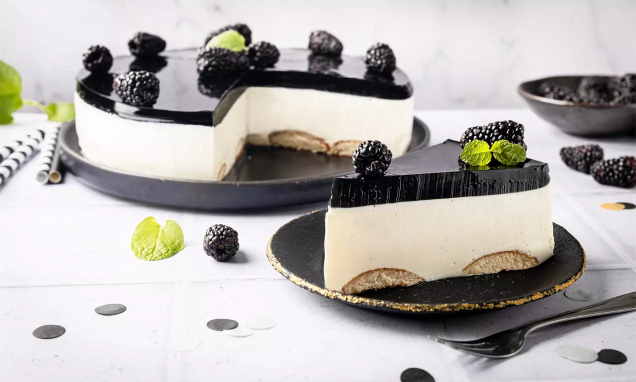 Czarno-biały sernik na zimno z galaretką i kremowym wnętrzem podanym na czarnym talerzu, ozdobiony świeżymi jeżynami i listkami mięty