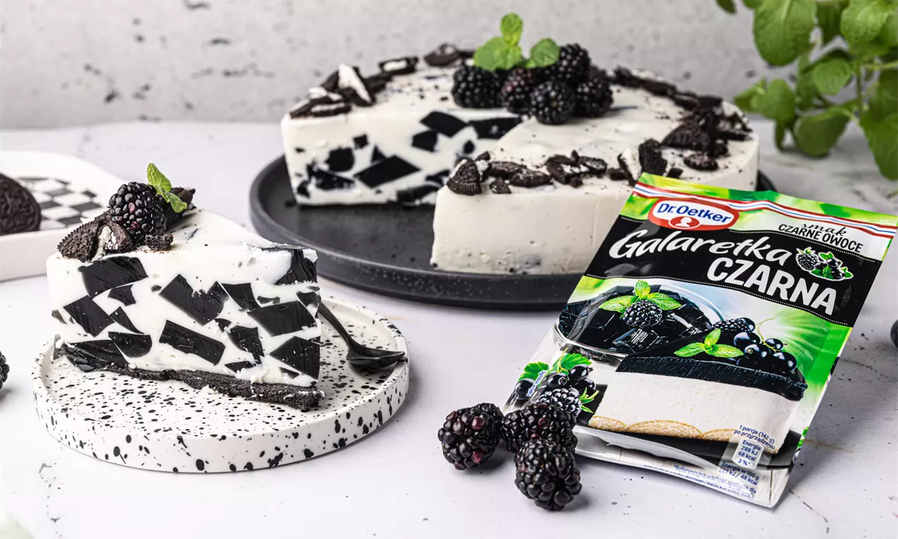 Sernik na zimno o kremowej białej strukturze z czarnymi geometrycznymi wzorami, udekorowany jeżynami, miętą i kawałkami ciasteczek na ceramicznym talerzu