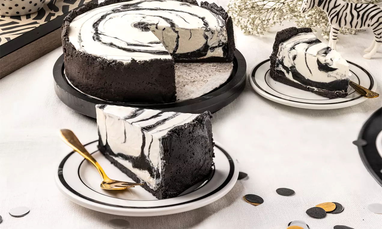 Sernik na zimno w paski czarno-białe z kremowym środkiem i spodem z ciemnych herbatników, podany na białych talerzach ze złotym widelcem