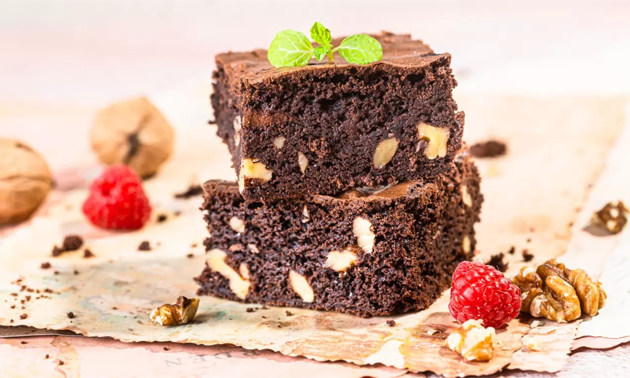 Kwadratowe kawałki ciemnego brownie z wyraźnymi kawałkami orzechów, ułożone na pergaminie, z malinami, orzechami włoskimi i gałązką mięty