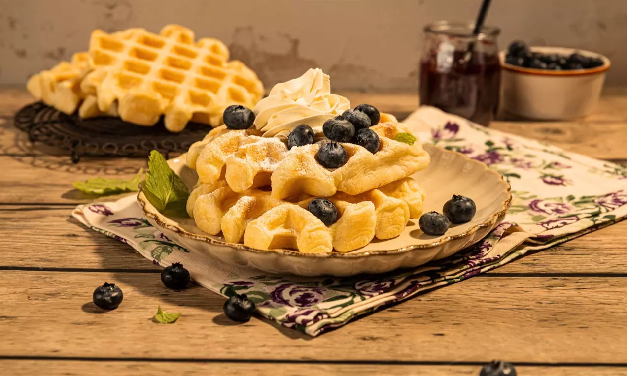 Gofry podane na talerzu z bitą śmietaną i borówkami, oprószone cukrem pudrem, na drewnianym stole.