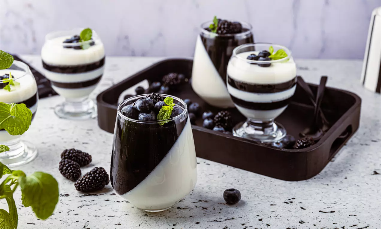 Warstwowe desery w kieliszkach z czarno-białymi paseczkami, udekorowane owocami jagodowymi i listkami mięty na marmurowej powierzchni