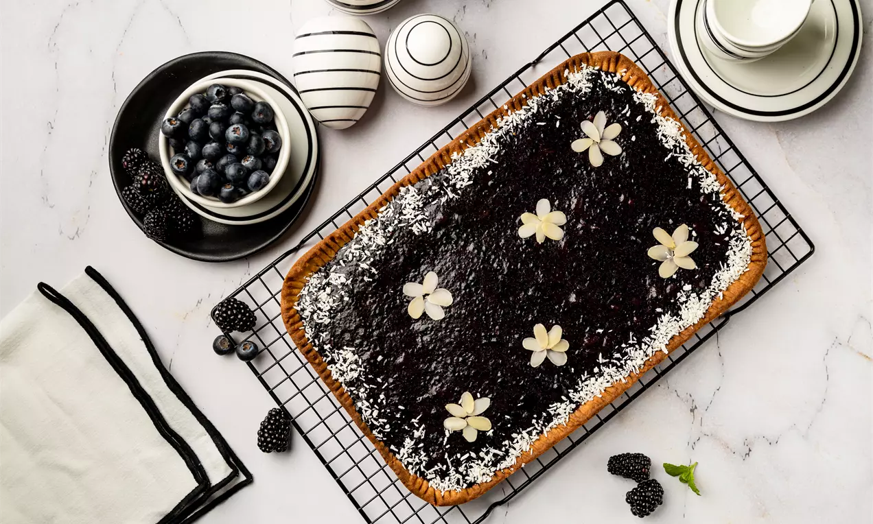 Mazurek prostokątny na złocistym spodzie, pokryty połyskującą czarną galaretką, ozdobiony cukrowymi kwiatkami i wiórkami kokosowymi