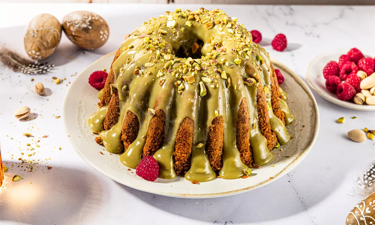 Babka w kształcie wieńca z zielonym pistacjowym lukrem, posypana kruszonymi pistacjami, podana na talerzu z malinami i orzechami