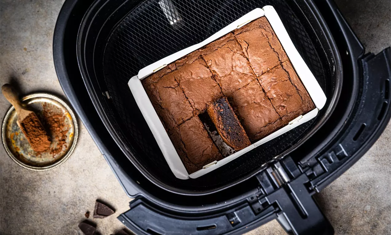 Kwadratowe brownie o wilgotnej, czekoladowej strukturze, upieczone w białej formie w koszu frytkownicy, obok kawałki czekolady i kakao na talerzyku